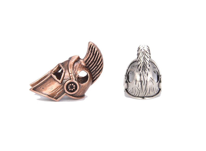 May include: Two metal helmet beads. One is bronze-colored with a detailed design, including a crest and circular accents. The other is silver-colored, also with a crest and detailed features. Both are on a white background.