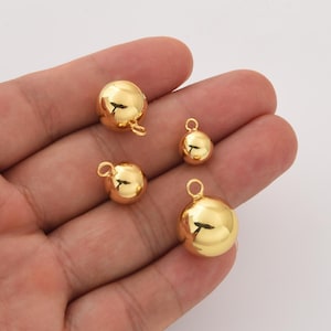 Puede incluir: Cuatro colgantes esféricos dorados de diferentes tamaños. Cada colgante tiene un lazo en la parte superior para sujetarlo a joyas u otras manualidades. Los colgantes son muy reflectantes, con una superficie lisa y pulida.