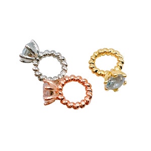 May include: Three engagement ring charms in silver, rose gold, and gold. Each ring features a clear, faceted gemstone and a twisted band. The charms are set against a white background.
