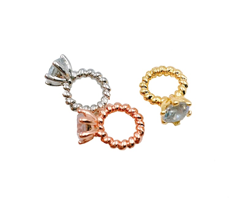 May include: Three engagement ring charms in silver, rose gold, and gold. Each ring features a clear, faceted gemstone and a twisted band. The charms are set against a white background.