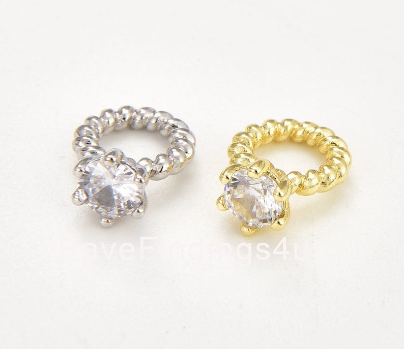 May include: Two ring charms, one silver and one gold, each with a clear, faceted gemstone. The ring bands have a twisted rope design. Ideal for jewellery making or craft projects.