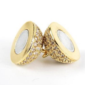 May include: Two gold-tone, oval-shaped,  pendants with a clear crystal-like stone design. The pendants have a smooth, shiny finish and a small loop on the top for attaching to a chain.