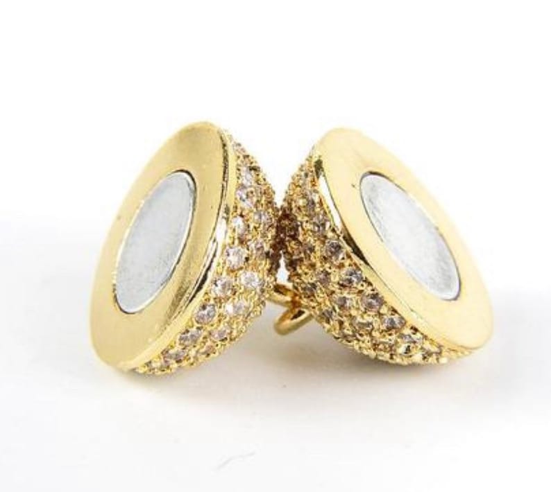 May include: Two gold-tone, oval-shaped,  pendants with a clear crystal-like stone design. The pendants have a smooth, shiny finish and a small loop on the top for attaching to a chain.