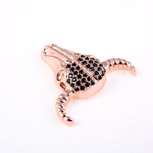 May include: A rose gold bull skull charm with black crystal accents. The charm is detailed with a textured surface and has a small hole for attaching to a bracelet or necklace.