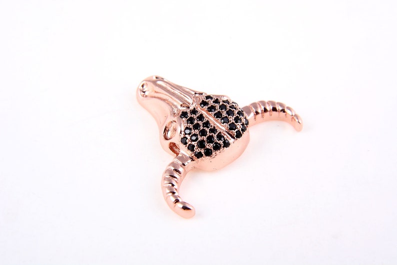 May include: A rose gold bull skull charm with black crystal accents. The charm is detailed with a textured surface and has a small hole for attaching to a bracelet or necklace.