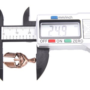 May include: A bronze-colored metal pendant, shaped like a helmet, is measured with a digital caliper. The digital display reads 24.9 mm. The caliper is black and silver, set against a white background.