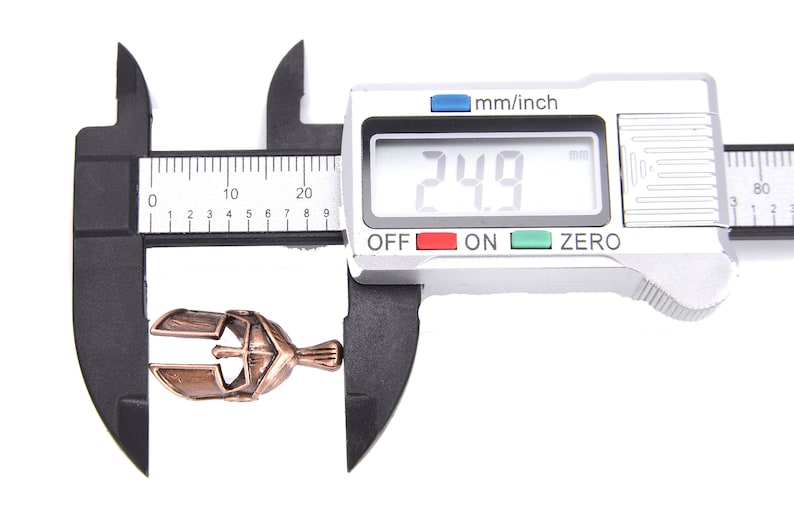 May include: A bronze-colored metal pendant, shaped like a helmet, is measured with a digital caliper. The digital display reads 24.9 mm. The caliper is black and silver, set against a white background.