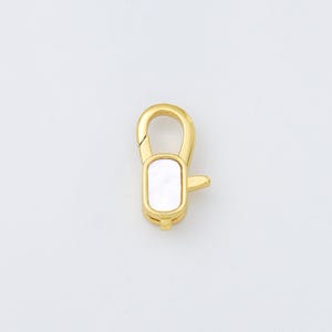 May include: A gold-colored lobster clasp with a rectangular, mother-of-pearl inlay. The clasp is designed with a curved top and a small lever for secure closure. The overall design is sleek and modern, suitable for jewelry making or crafting.