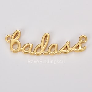 May include: Gold-colored metal charm with the word "badass" written in a cursive font.