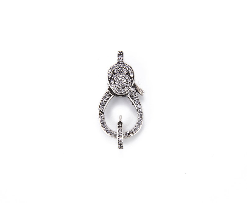 May include: Silver-toned clasp with a clear crystal design. The clasp is oval-shaped and has a small, clear crystal in the center.