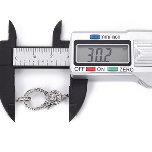 May include: A silver-toned clasp with clear sparkling stones is measured with a digital caliper. The caliper reads 30.2 mm.