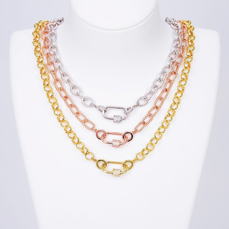 K&ouml;nnte beinhalten: Drei goldene Kettenhalsb&auml;nder mit einem Verschluss. Die Ketten sind &uuml;bereinander gelegt. Die oberste Kette ist silber, die mittlere Kette ist ros&eacute;gold und die unterste Kette ist gold. Jeder Verschluss hat ein kleines Quadrat aus klaren Zirkonia-Steinen.