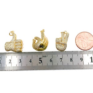 May include: Four gold-tone charms with clear rhinestones. The charms are shaped like a thumbs up, a peace sign, and two different versions of an OK sign. The charms are lined up on a ruler with a US quarter for size comparison.