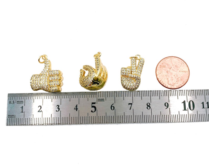 May include: Four gold-tone charms with clear rhinestones. The charms are shaped like a thumbs up, a peace sign, and two different versions of an OK sign. The charms are lined up on a ruler with a US quarter for size comparison.