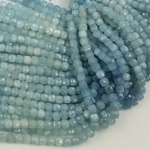 May include: A close-up of a bundle of aquamarine gemstone beads. The beads are square-shaped with faceted surfaces, displaying a range of light blue hues. The beads are strung together, ready for jewelry making.