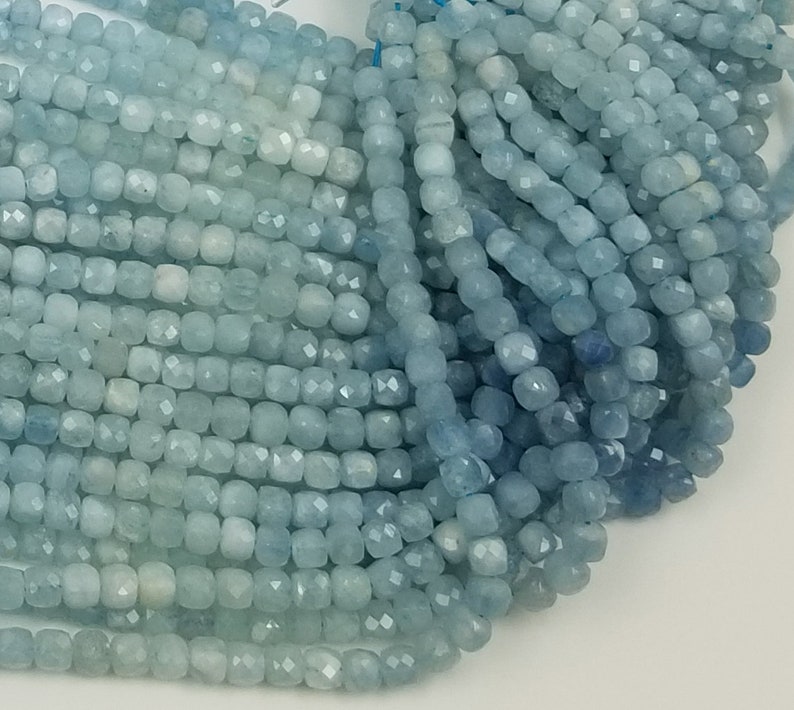 May include: A close-up of a bundle of aquamarine gemstone beads. The beads are square-shaped with faceted surfaces, displaying a range of light blue hues. The beads are strung together, ready for jewelry making.