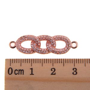 May include: Rose gold-colored jewelry connector with three oval links, each encrusted with small, clear crystals. The connector has small loops on each end for attachment. A wooden ruler with centimeter markings is in the foreground.