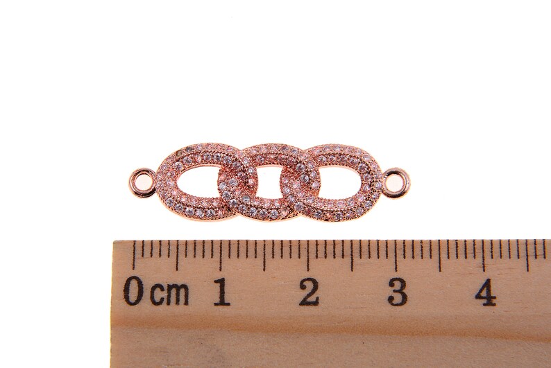 May include: Rose gold-colored jewelry connector with three oval links, each encrusted with small, clear crystals. The connector has small loops on each end for attachment. A wooden ruler with centimeter markings is in the foreground.
