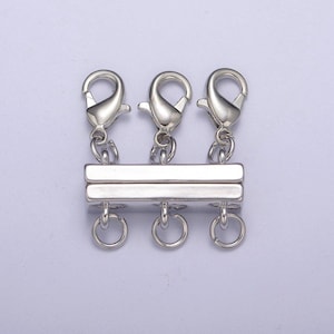 May include: A silver-toned magnetic jewelry clasp featuring three lobster claw clasps attached to the top bar and three jump rings attached to the bottom bar, designed for connecting multiple strands of necklaces or bracelets securely.