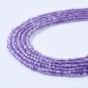 May include: Close-up of several strands of amethyst gemstone beads. The beads are a light purple color and appear to be faceted, catching the light. The strands are arranged in a curved pattern against a white background, suggesting they are part of a necklace or bracelet.