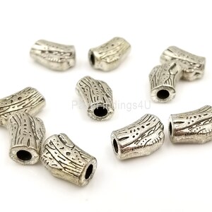 May include: A collection of silver-colored metal beads. Each bead is barrel-shaped with a textured, wood-grain design. The beads have a hole running through the center, and are scattered on a white surface.