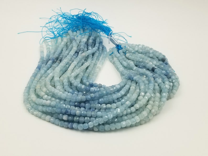 May include: Strands of faceted aquamarine beads in varying shades of blue and white. The beads are small, square-shaped, and strung together. A blue tassel is attached to the top of the strands. The beads are suitable for jewelry making.