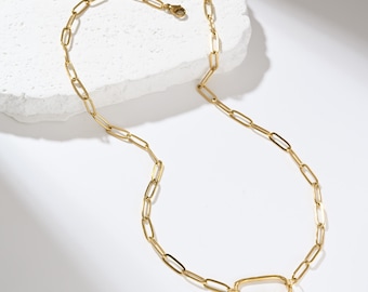 May include: A gold-colored chain necklace with an elongated oval clasp. The necklace features a series of rectangular links connected to a lobster clasp. The clasp has a small, decorative element. The jewelry is displayed on a white surface.