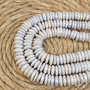 Puede incluir: Una hebra de cuentas de concha blanca iridiscente, cada cuenta es de forma ovalada plana con una superficie lisa y pulida. Las cuentas están dispuestas en un patrón circular sobre una superficie tejida.
