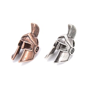 May include: Two miniature Spartan helmets, one in a bronze finish and the other in silver. Each helmet features a detailed crest and cheek guards, with a circular design on the side. The helmets are displayed against a white background.