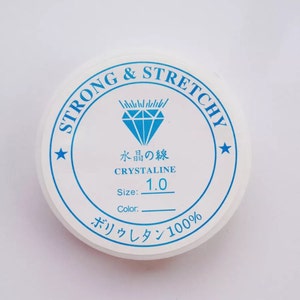 Puede incluir: Un recipiente redondo blanco con texto azul y un gr&aacute;fico de diamante azul. El texto dice "STRONG & STRETCHY", "水晶の線", "CRYSTALINE", "1.0", "Size:", "Color:" y "ボリウレタン100%".