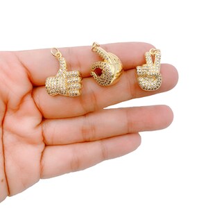 May include: Three gold-tone charms in the shape of hands making different gestures. The charms are covered in clear sparkling rhinestones. The charms are on a hand.