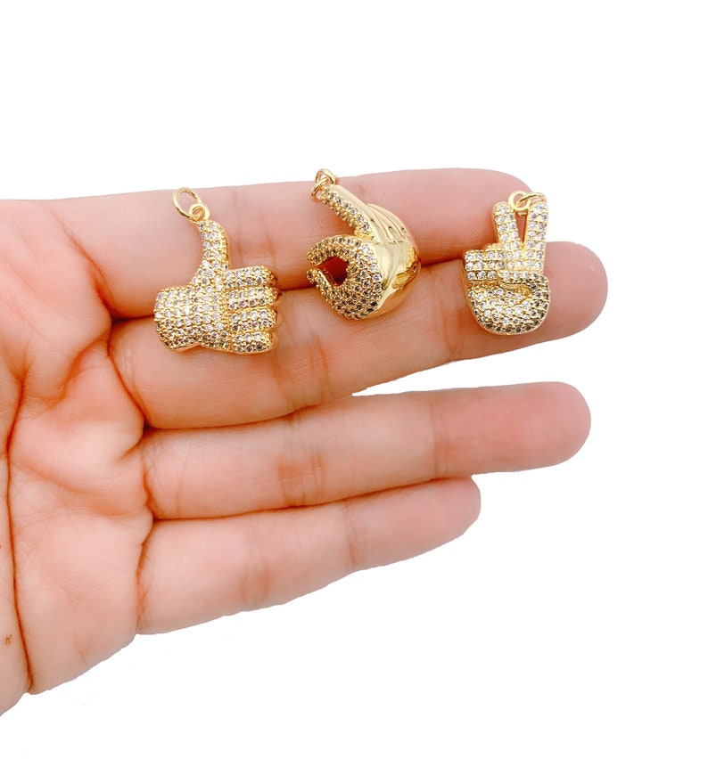 May include: Three gold-tone charms in the shape of hands making different gestures. The charms are covered in clear sparkling rhinestones. The charms are on a hand.
