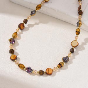 Puede incluir: Un collar de cuentas con una mezcla de cuentas marrones, ámbar y moradas, junto con pequeñas cuentas doradas. El collar tiene un diseño elegante, adecuado para el uso diario o para ocasiones especiales. Las cuentas se miden en centímetros.