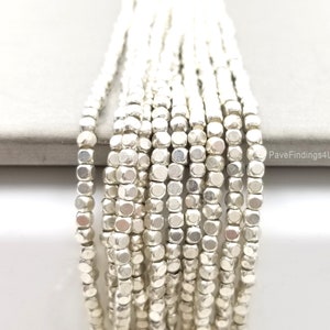 Puede incluir: Hebras de cuentas plateadas en forma de cubo. Las cuentas están ensartadas, creando múltiples líneas verticales. Las cuentas tienen un brillo metálico y una forma ligeramente irregular. El fondo es una superficie gris claro.