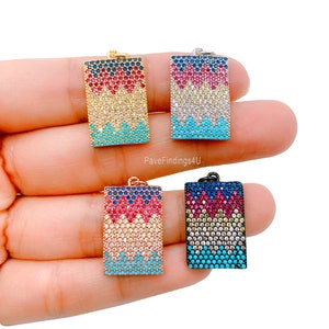 Puede incluir: Cuatro colgantes rectangulares con un patrón geométrico de piedras de circonita cúbica turquesa, blancas, rosas y azules. Los colgantes se muestran en una mano, con los colgantes dorados, plateados, de oro rosa y negros visibles.