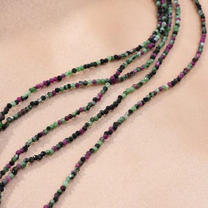 May include: A strand of green, black, and pink gemstone beads. The beads are small and round, and they are strung on a clear thread. The beads are arranged in a repeating pattern.