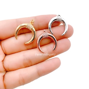 May include: Three crescent moon pendants in gold, rose gold, and silver. Each pendant features a row of small, clear stones at the top. The pendants are displayed against a light background.