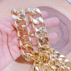 May include: A gold-colored chain necklace with large, chunky links. The necklace is held in a person's hand, showcasing the shiny, reflective surface of the metal. The chain is a bold statement piece.