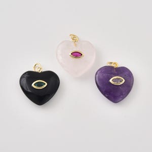 May include: Three heart-shaped pendants featuring a gold-tone bail at the top. One is black with a green marquise-cut stone, one is pale pink with a red marquise-cut stone, and one is purple with a clear marquise-cut stone set in the center.