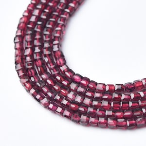 May include: Close-up of several strands of faceted, deep red garnet beads. The beads are small, square-cut, and strung together, creating a rich, textured appearance. The color is a vibrant, translucent red, with hints of darker shades.