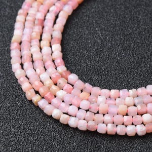 May include: Strands of faceted, light pink opal beads. The beads are cube-shaped and strung together, displaying variations in color from pale pink to peach. The background is a textured black surface.