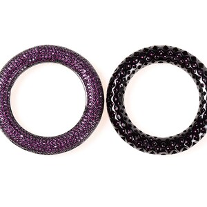 May include: Two circular bangles. One is covered in small, sparkling purple gems. The other is black with a geometric pattern and small purple accents. Both are set against a white background.