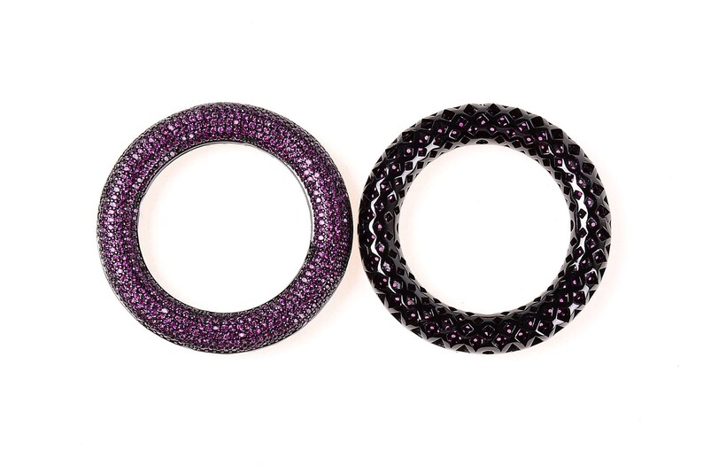 May include: Two circular bangles. One is covered in small, sparkling purple gems. The other is black with a geometric pattern and small purple accents. Both are set against a white background.