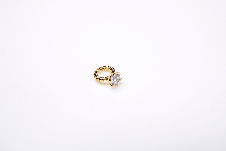 May include: A gold-coloured ring featuring a large, clear, round gemstone. The ring band has a textured, twisted design. The ring is set against a plain white background.