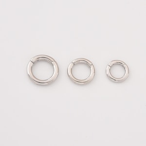 May include: Three silver hinged rings of different sizes are displayed on a white background. The rings are circular with a smooth, metallic finish. The sizes vary, presenting a set of body jewellery.