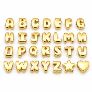Peut inclure: Une collection de perles alphabet en métal doré disposées sur quatre rangées. L'ensemble comprend toutes les lettres de A à Z, ainsi qu'une forme d'étoile à cinq branches et une forme de cœur. Chaque pièce a une surface lisse, réfléchissante et arrondie.
