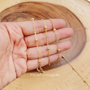 May include: Three gold chains with small round beads. The chains are different lengths and are displayed on a hand.