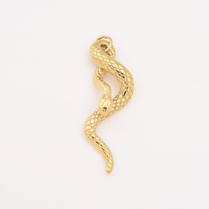 May include: A gold-coloured snake pendant with a textured, scale-like pattern. The snake is coiled in a stylised, serpentine shape, with a detailed head and a loop for hanging. The pendant is set against a white background.