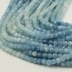 May include: Strands of aquamarine gemstone beads in varying shades of blue. The beads are small, square-cut, and faceted, creating a sparkling effect. The arrangement suggests a jewelry-making supply or finished necklace.