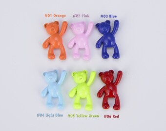 May include: Six small plastic teddy bear figurines in various colors: orange, pink, blue, light blue, yellow green, and red. Each bear is standing with one arm raised.